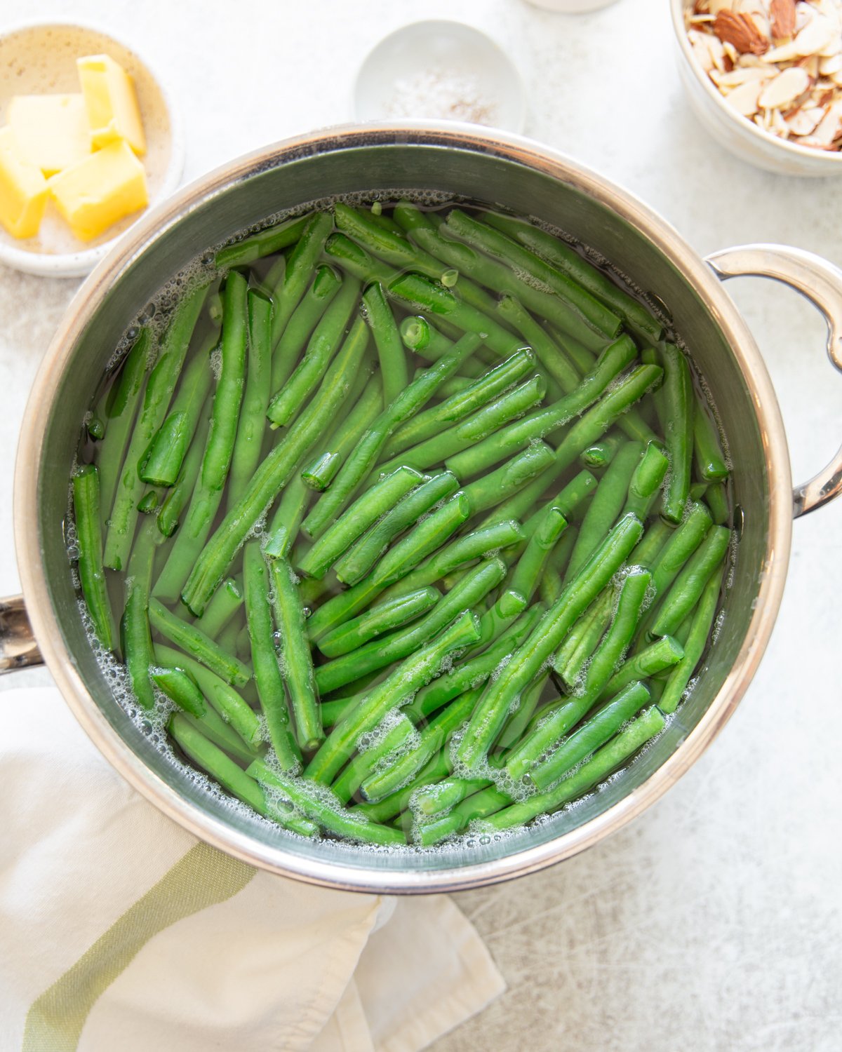 A pot filled with green beans boiling in water sits on a white surface. Nearby are small bowls with butter, sliced almonds, and salt. A white towel with a green stripe is draped next to the pot.