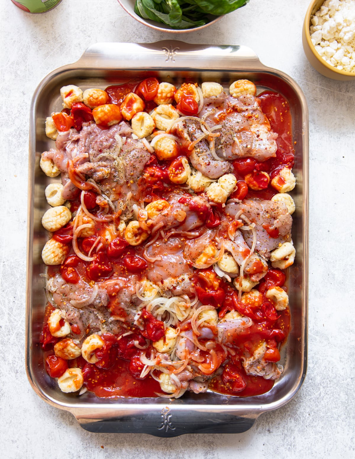 A metal baking pan filled with raw chicken thighs, gnocchi, halved cherry tomatoes, sliced onions, tomato sauce, and Italian seasonings, ready to be baked. Fresh basil leaves and cheese are in bowls nearby.