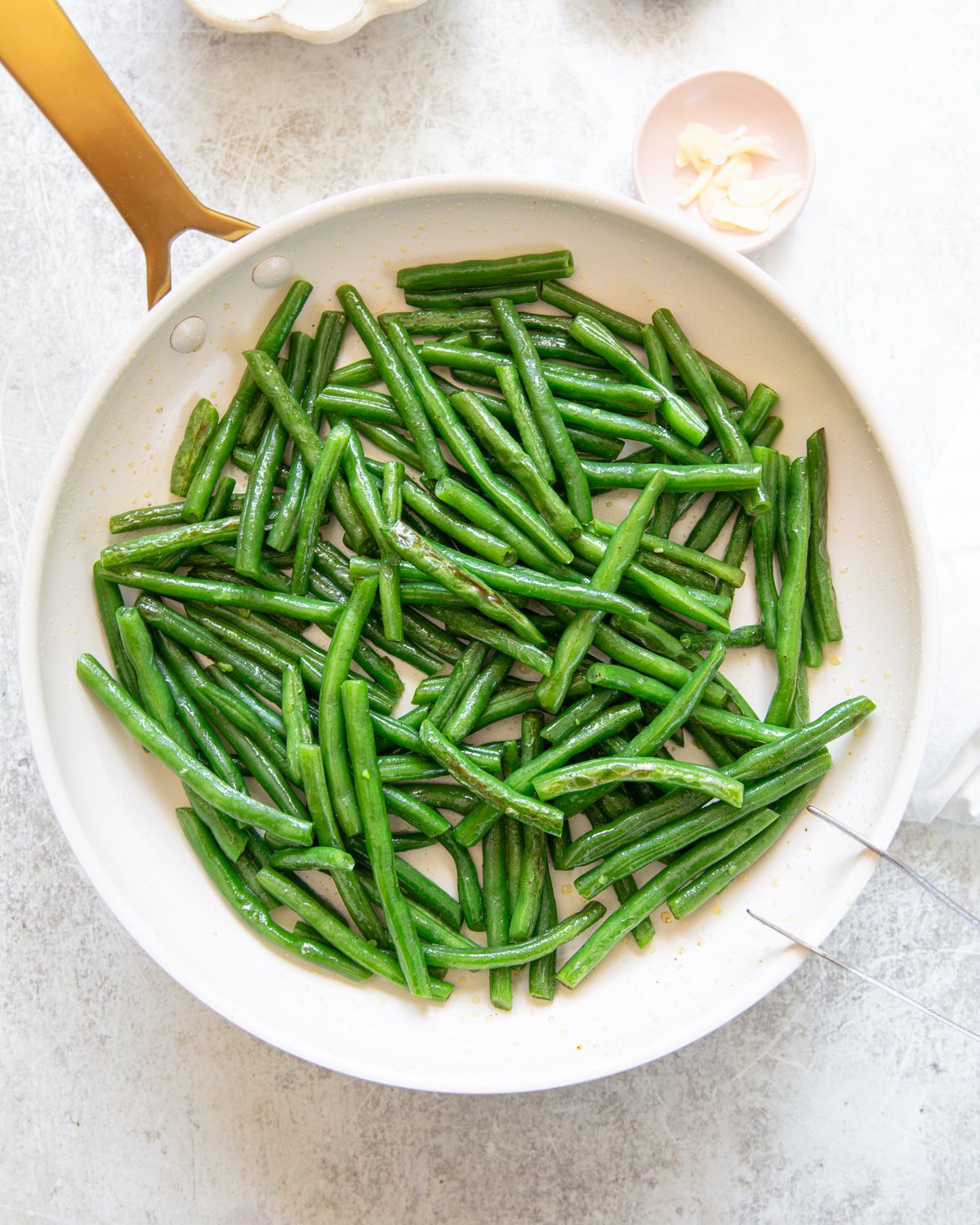 A white skillet with a golden handle contains freshly sautéed green beans. The skillet sits on a light-colored surface, with a small dish of garlic slices visible in the background.
