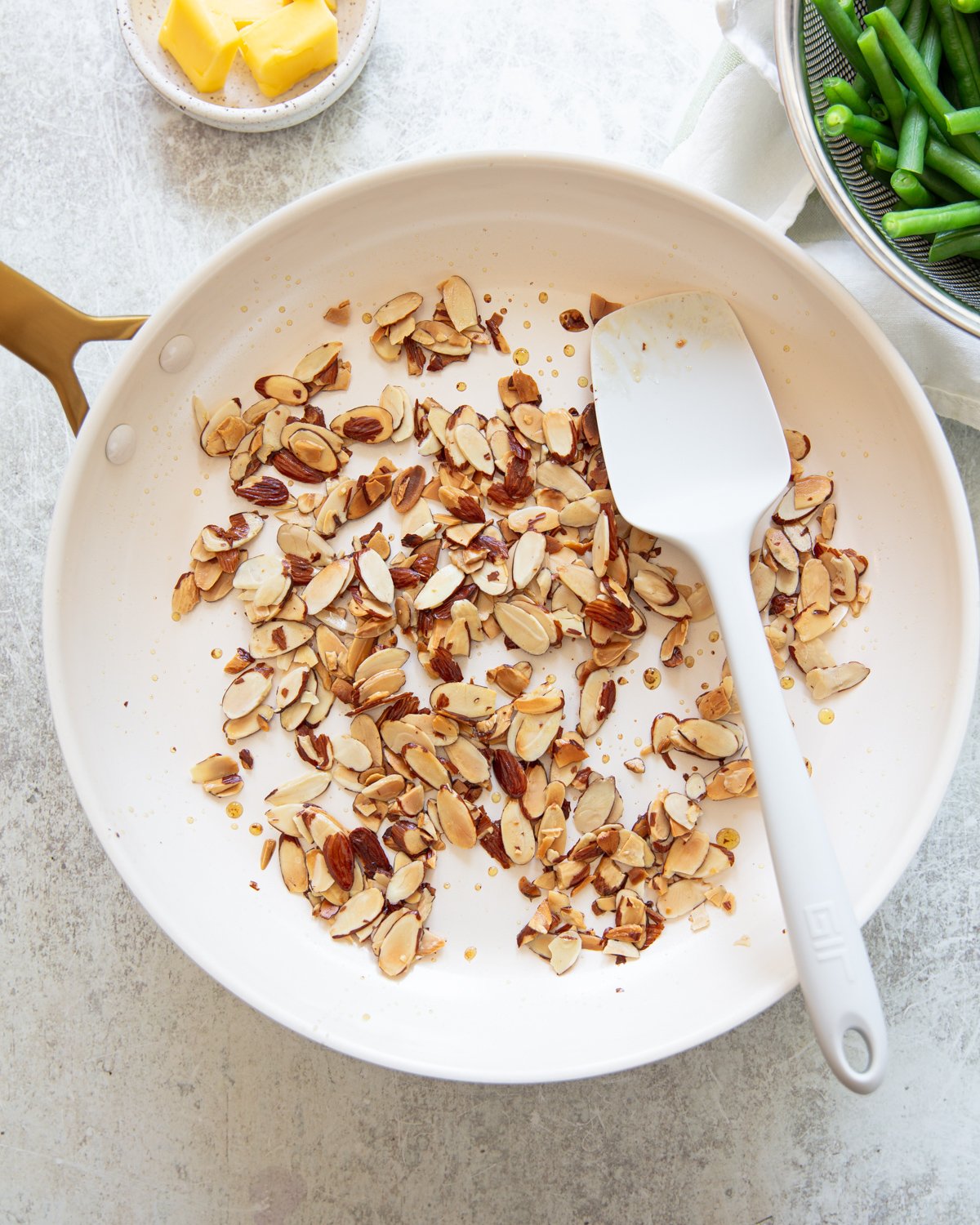 A white skillet contains toasted sliced almonds, stirred with a white spatula. Nearby are a small dish of butter and a bowl of green beans.