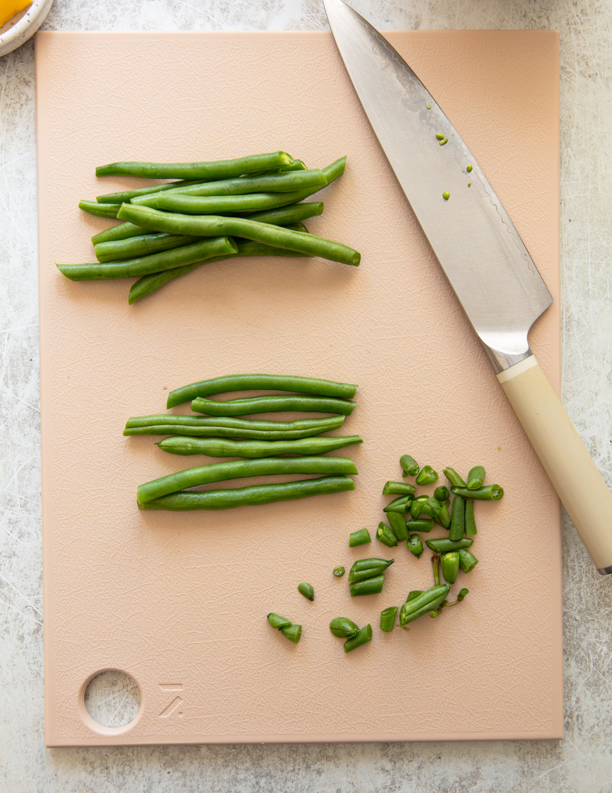 A beige cutting board with whole green beans, sliced green beans, chopped green bean ends, and a large knife with a light handle on a pale surface.