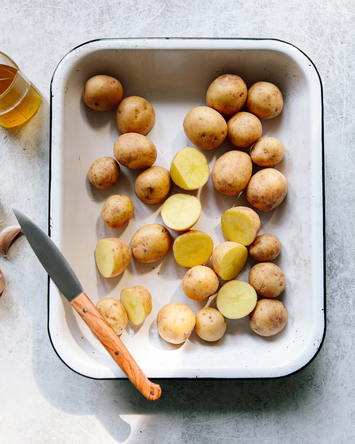 A white enamel baking dish filled with whole and halved small potatoes sits on a light surface. A knife with a wooden handle rests on the edge of the dish, and a glass filled with oil is nearby.