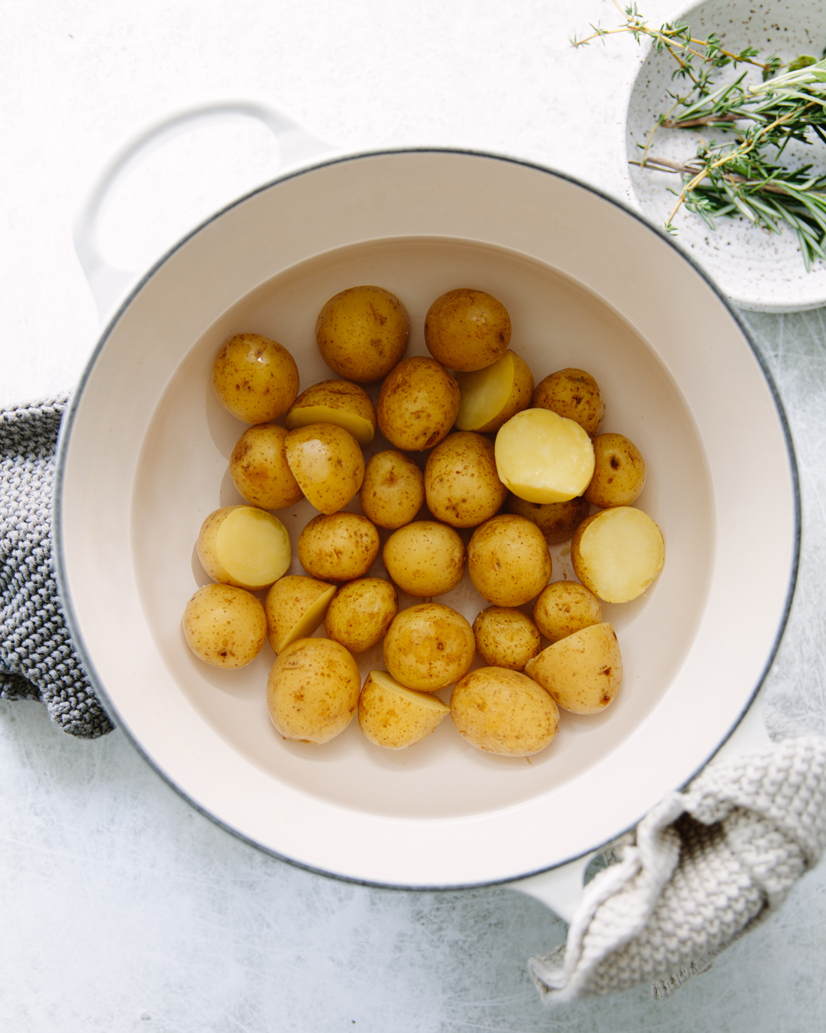 A white pot filled with water and small yellow potatoes, some whole and some halved, sits on a white surface. Fresh herbs and a gray cloth are nearby.
