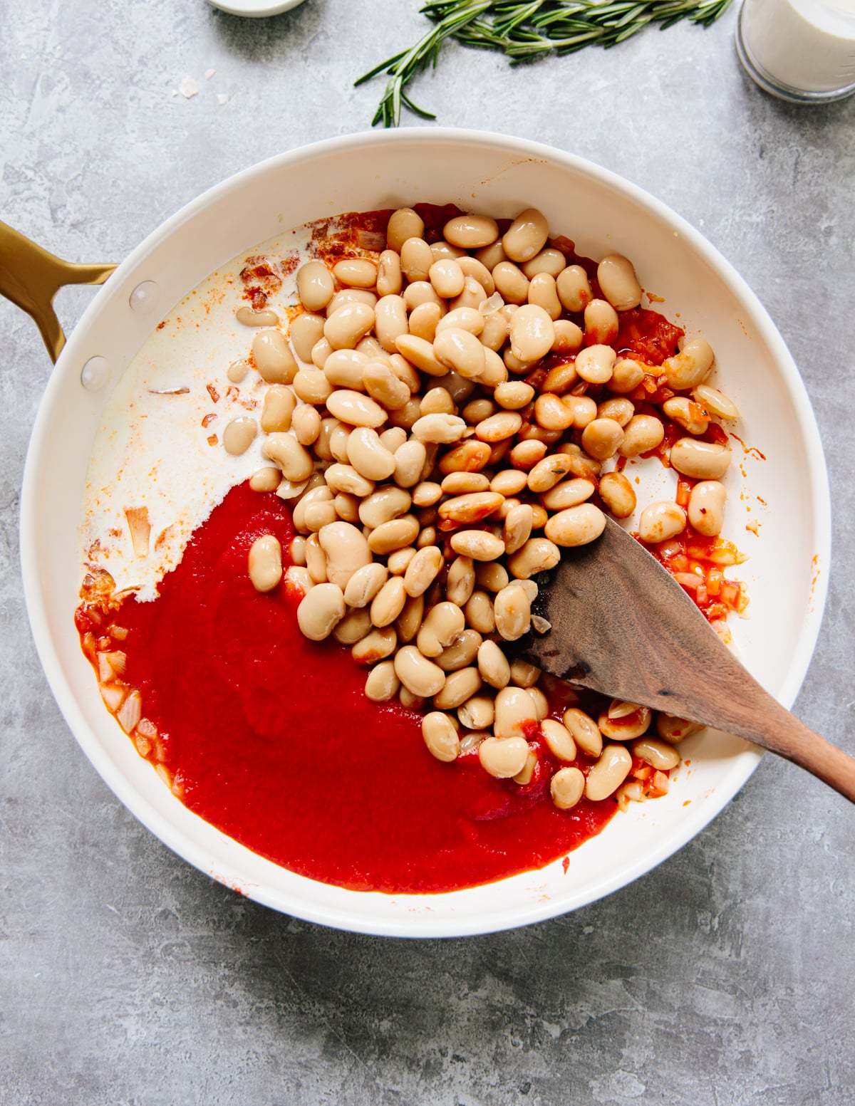 A white skillet with cooked onions, white beans, and tomato sauce being stirred with a wooden spoon, on a gray countertop with a sprig of rosemary and a small bowl nearby.
