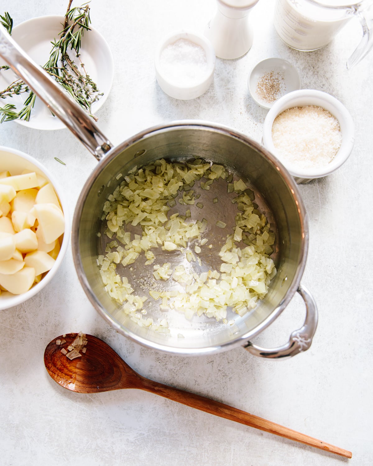 Chopped onions sautéing in a pot, surrounded by bowls of cut potatoes, grated cheese, salt, pepper, cream, and rosemary, with a wooden spoon resting nearby on a light countertop.