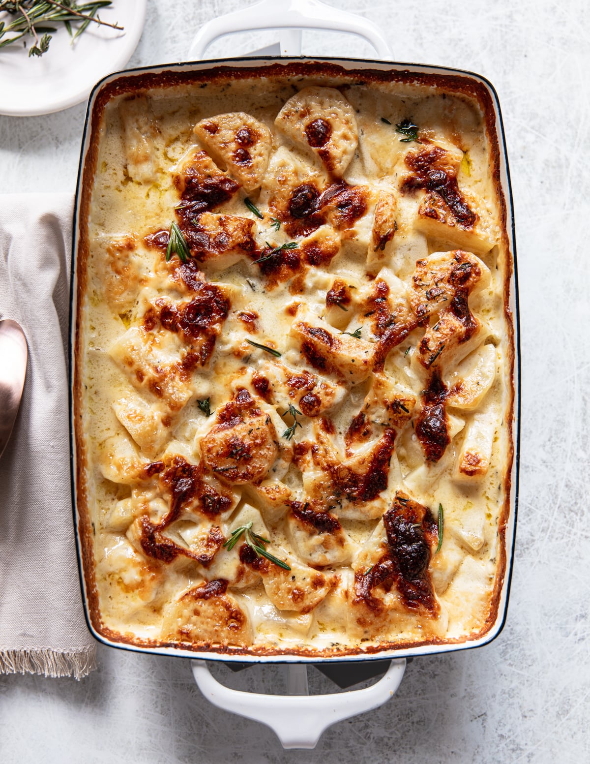 A white baking dish filled with golden-brown, creamy potato gratin garnished with herbs, resting on a light surface next to a beige napkin and a spoon.
