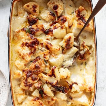 A baked casserole dish filled with golden-brown, cheesy scalloped potatoes, topped with herbs, and a serving spoon scooping out a portion.