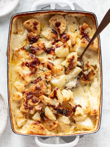 A baked casserole dish filled with golden-brown, cheesy scalloped potatoes, topped with herbs, and a serving spoon scooping out a portion.