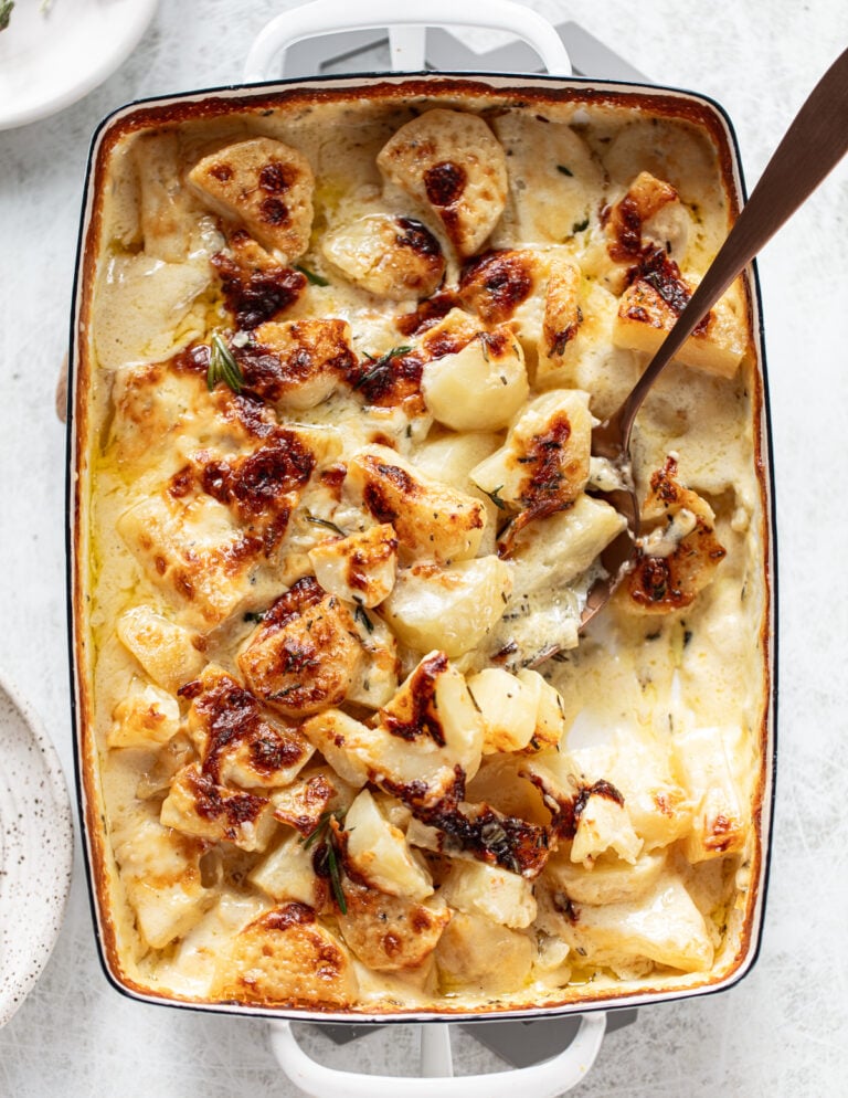 A baked casserole dish filled with golden-brown, cheesy scalloped potatoes, topped with herbs, and a serving spoon scooping out a portion.
