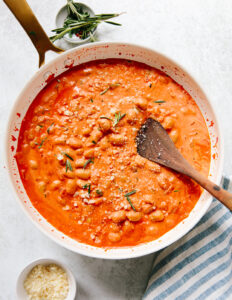 A skillet filled with creamy tomato and white bean stew, topped with grated cheese and rosemary, with a wooden spoon resting in it. There’s a striped towel, a bowl of grated cheese, and herbs nearby.