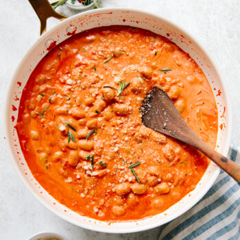 A skillet filled with creamy tomato and white bean stew, topped with grated cheese and rosemary, with a wooden spoon resting in it. There’s a striped towel, a bowl of grated cheese, and herbs nearby.