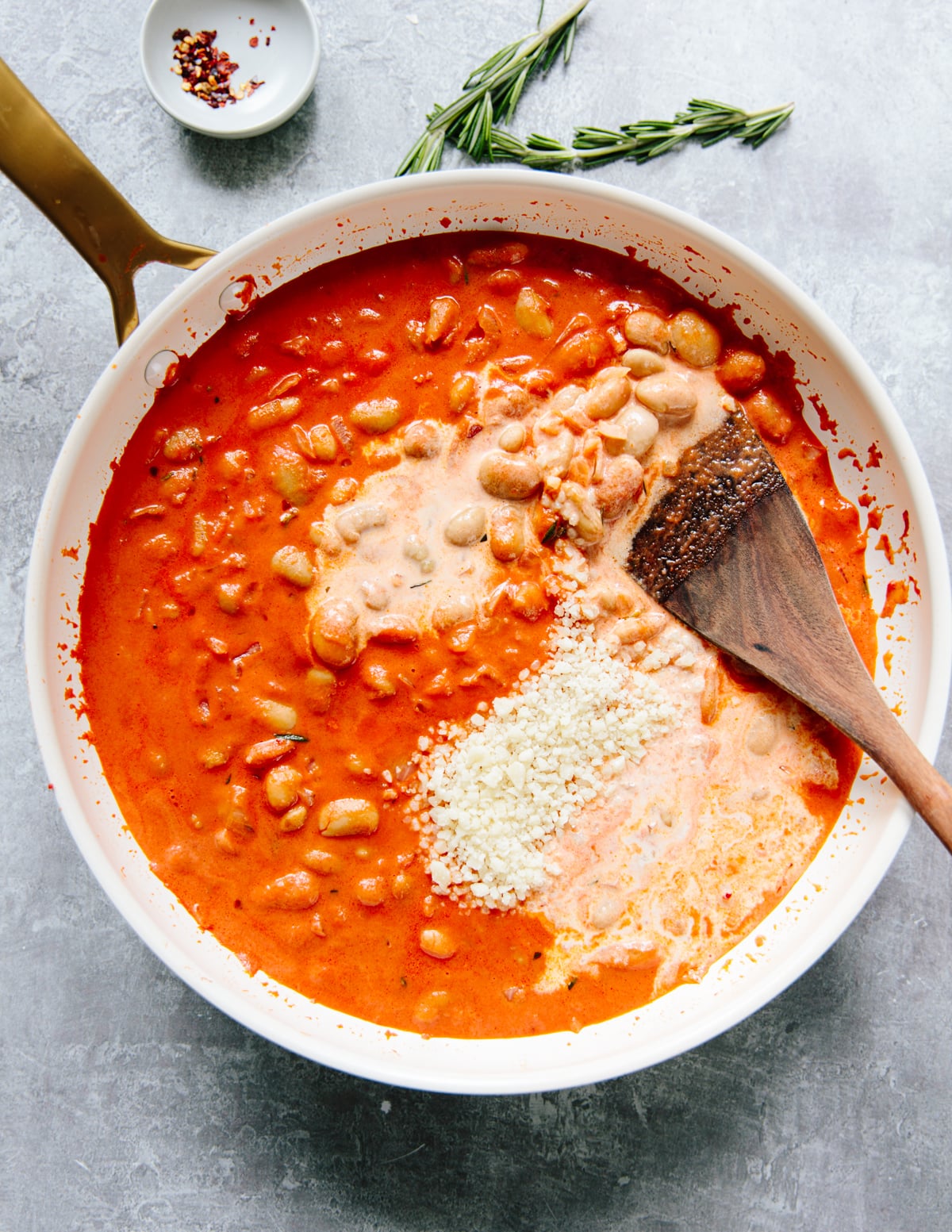 A white skillet with a creamy tomato sauce, cooked beans, and grated cheese being stirred with a wooden spoon. Fresh rosemary and a small dish of chili flakes sit nearby on a gray surface.