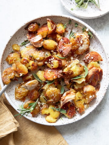 A plate of crispy smashed potatoes garnished with fresh rosemary, sage, roasted garlic cloves, and grated cheese. The dish sits on a white speckled plate with a beige napkin beside it.