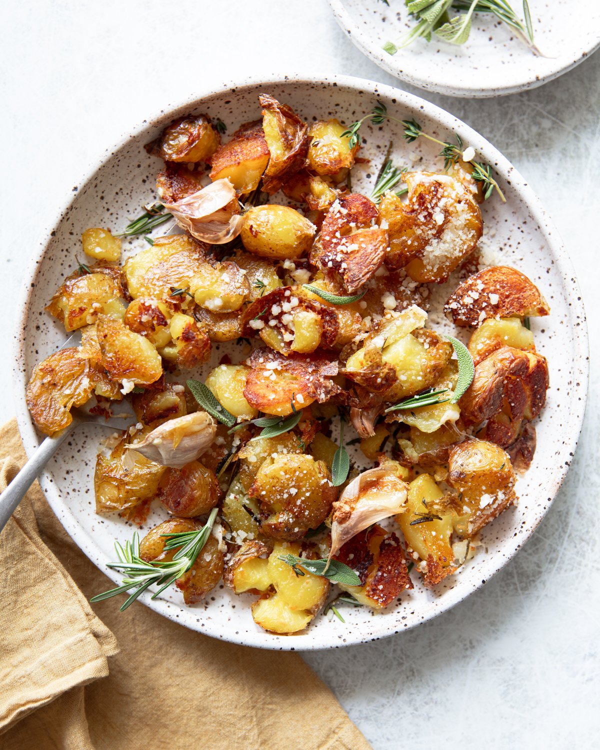 A plate of crispy smashed potatoes garnished with fresh rosemary, sage, roasted garlic cloves, and grated cheese. The dish sits on a white speckled plate with a beige napkin beside it.