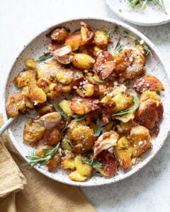 A plate of crispy smashed potatoes garnished with fresh rosemary, grated cheese, and roasted garlic cloves, served on a speckled ceramic dish with a napkin and herbs on the side.