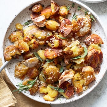 A plate of crispy smashed potatoes garnished with fresh rosemary, grated cheese, and roasted garlic cloves, served on a speckled ceramic dish with a napkin and herbs on the side.