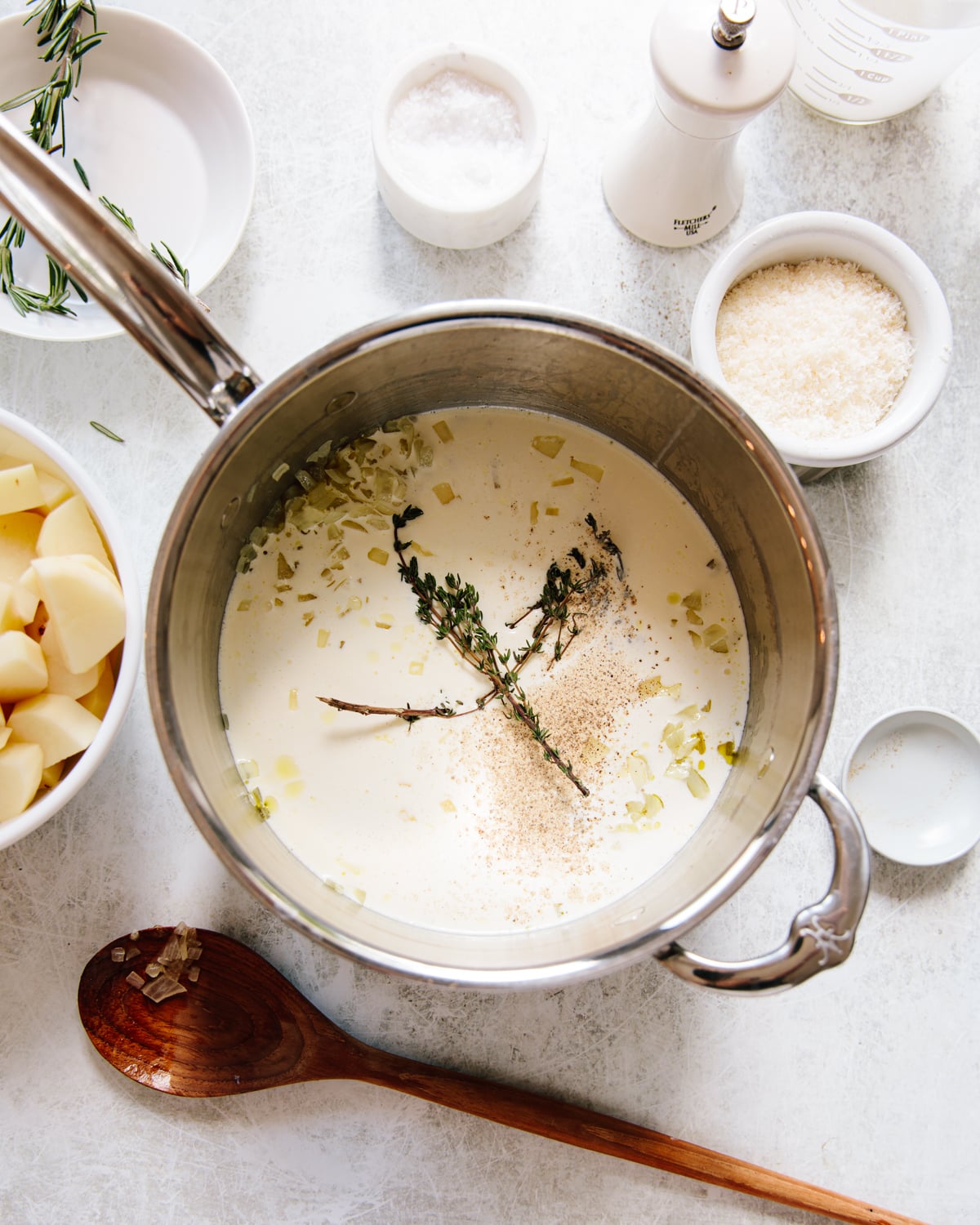 A saucepan filled with cream, thyme sprigs, and black pepper sits on a white surface, surrounded by bowls of salt, grated cheese, chopped potatoes, a wooden spoon, and a pepper grinder.