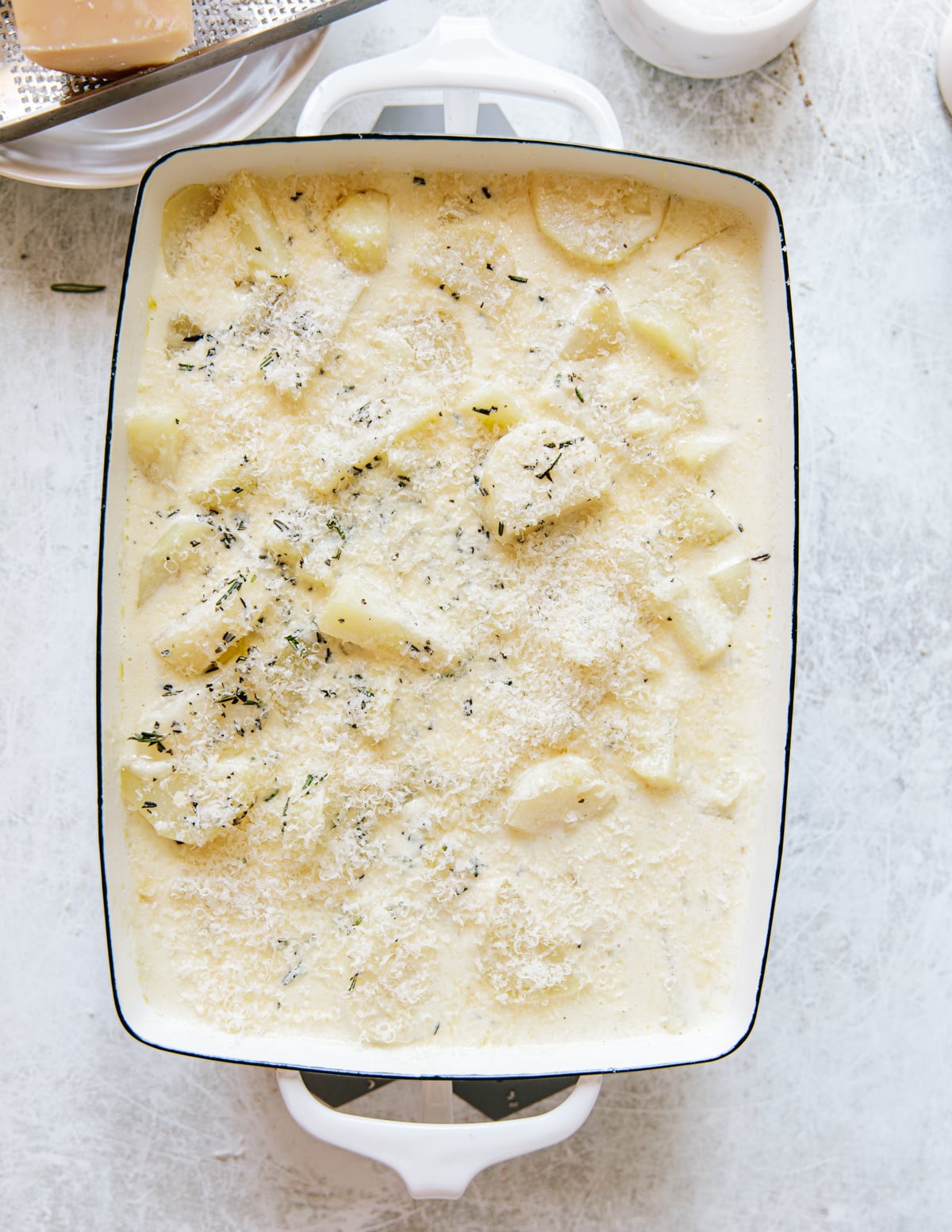 A white baking dish filled with sliced potatoes in a creamy sauce, topped with grated cheese and sprinkled with herbs, ready to be baked. The dish is on a light-colored surface.