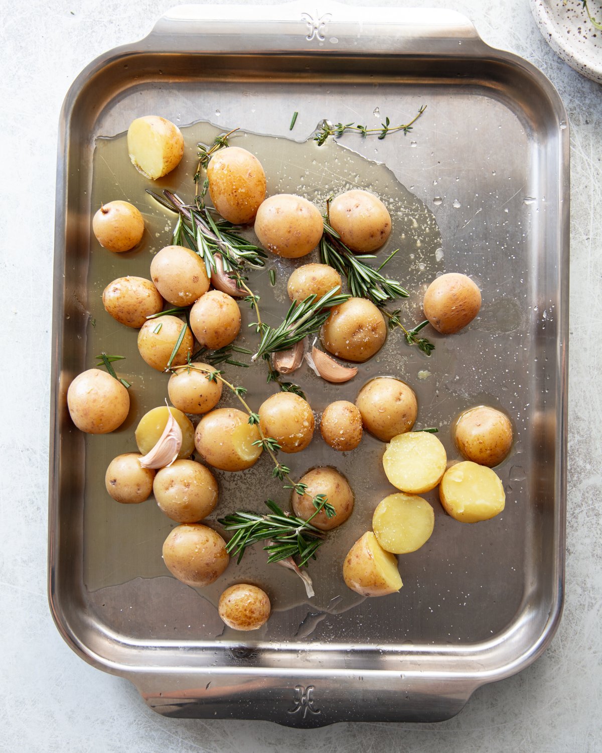 A metal baking tray with small whole potatoes, some halved, scattered with fresh rosemary sprigs, garlic cloves, and a drizzle of olive oil.