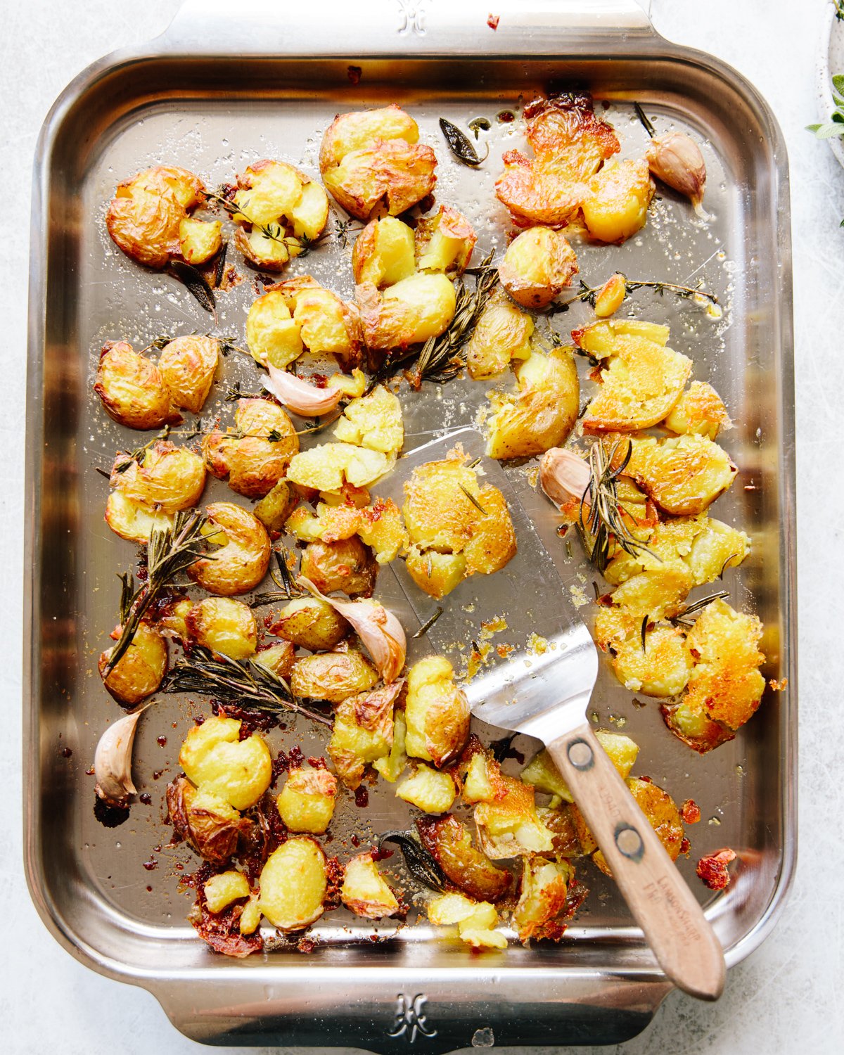 A metal baking tray filled with baked smashed potatoes, roasted garlic cloves, and sprigs of rosemary. A spatula with a wooden handle rests on the tray among the golden, slightly browned potatoes.