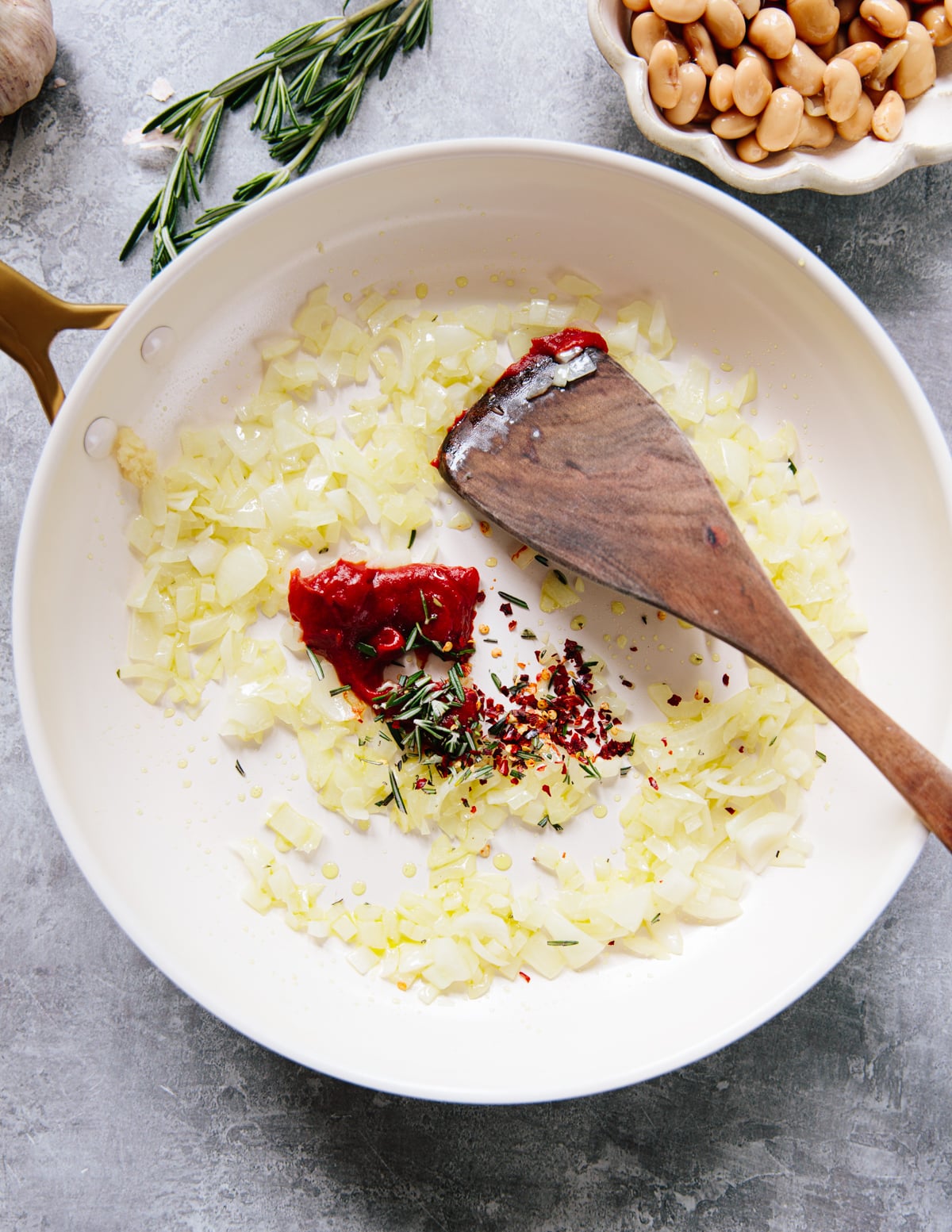 Chopped onions, tomato paste, rosemary, red pepper flakes, and olive oil are being sautéed in a white skillet with a wooden spatula. A bowl of white beans and sprigs of rosemary are nearby.