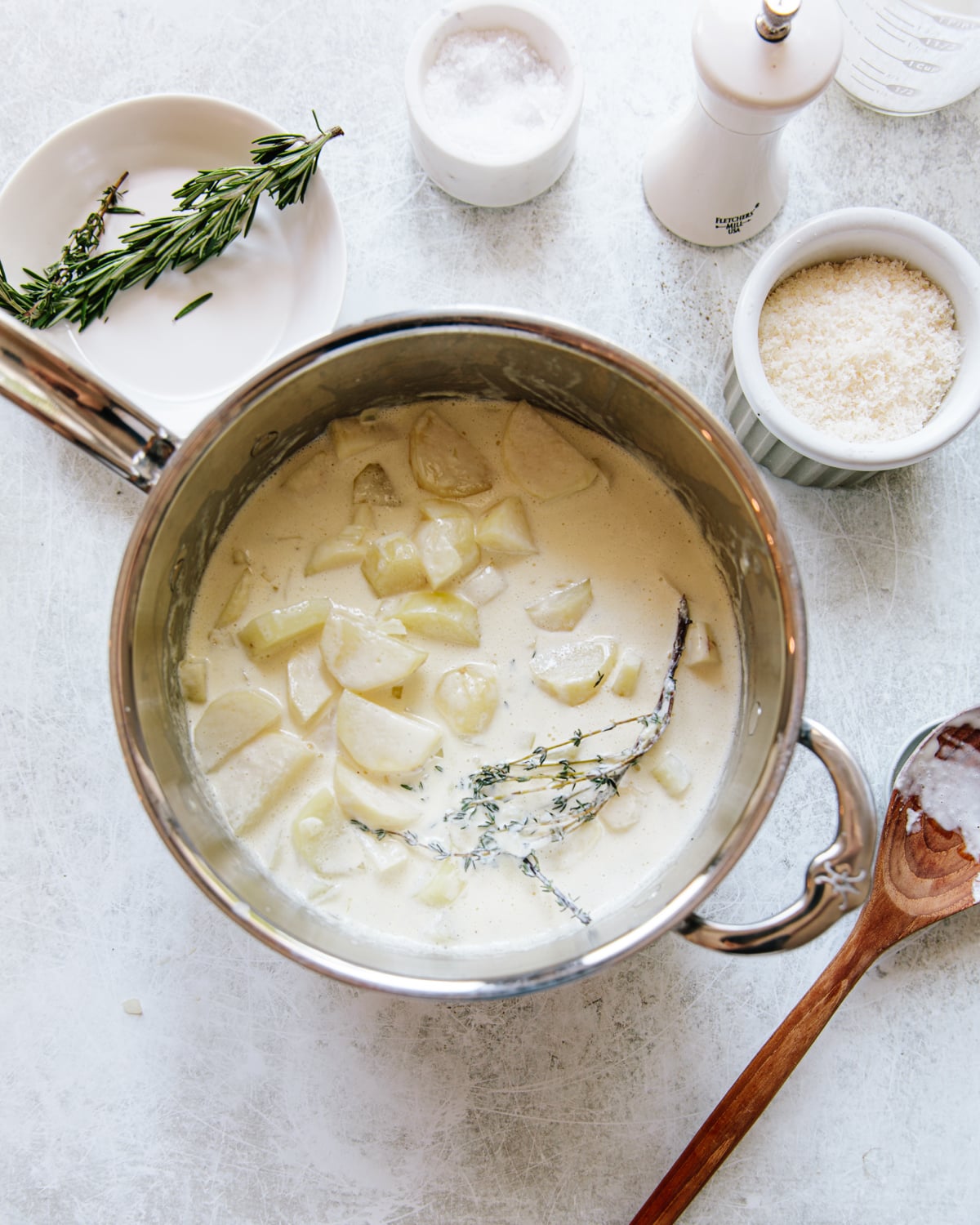 A saucepan filled with sliced potatoes, cream, and herbs sits on a light countertop. Nearby are a plate with rosemary, a bowl of grated cheese, a bowl of salt, a pepper grinder, and a wooden spoon.