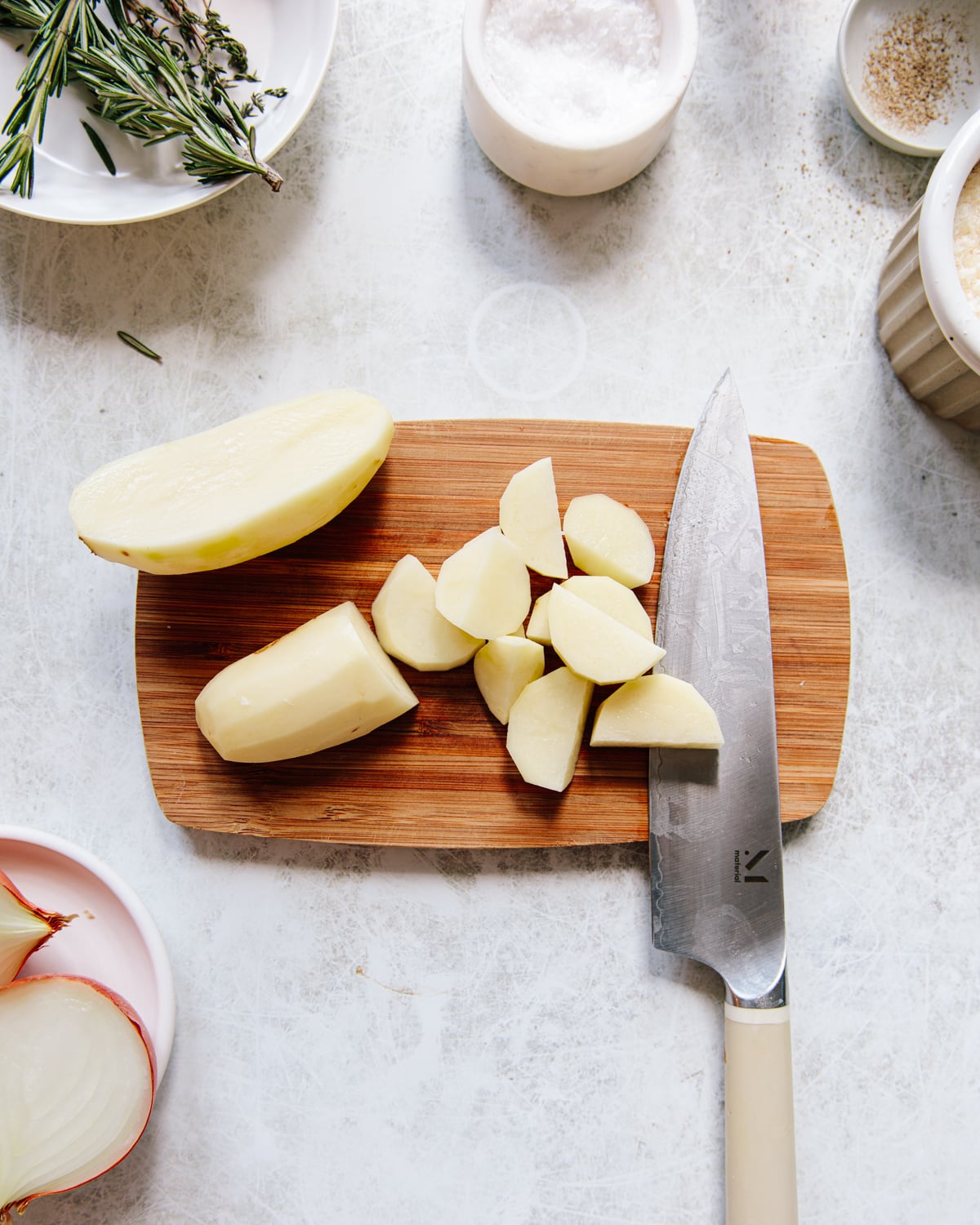 A wooden cutting board with a peeled, halved and chopped potato, a chef’s knife, and bowls of salt, herbs, and sliced onions on a light countertop.