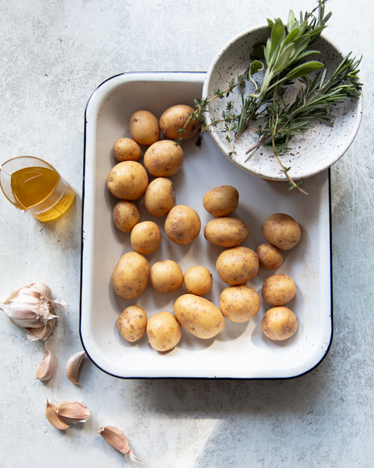 A white tray with small yellow potatoes, next to garlic cloves, a glass of olive oil, and a bowl containing fresh herbs on a light surface.