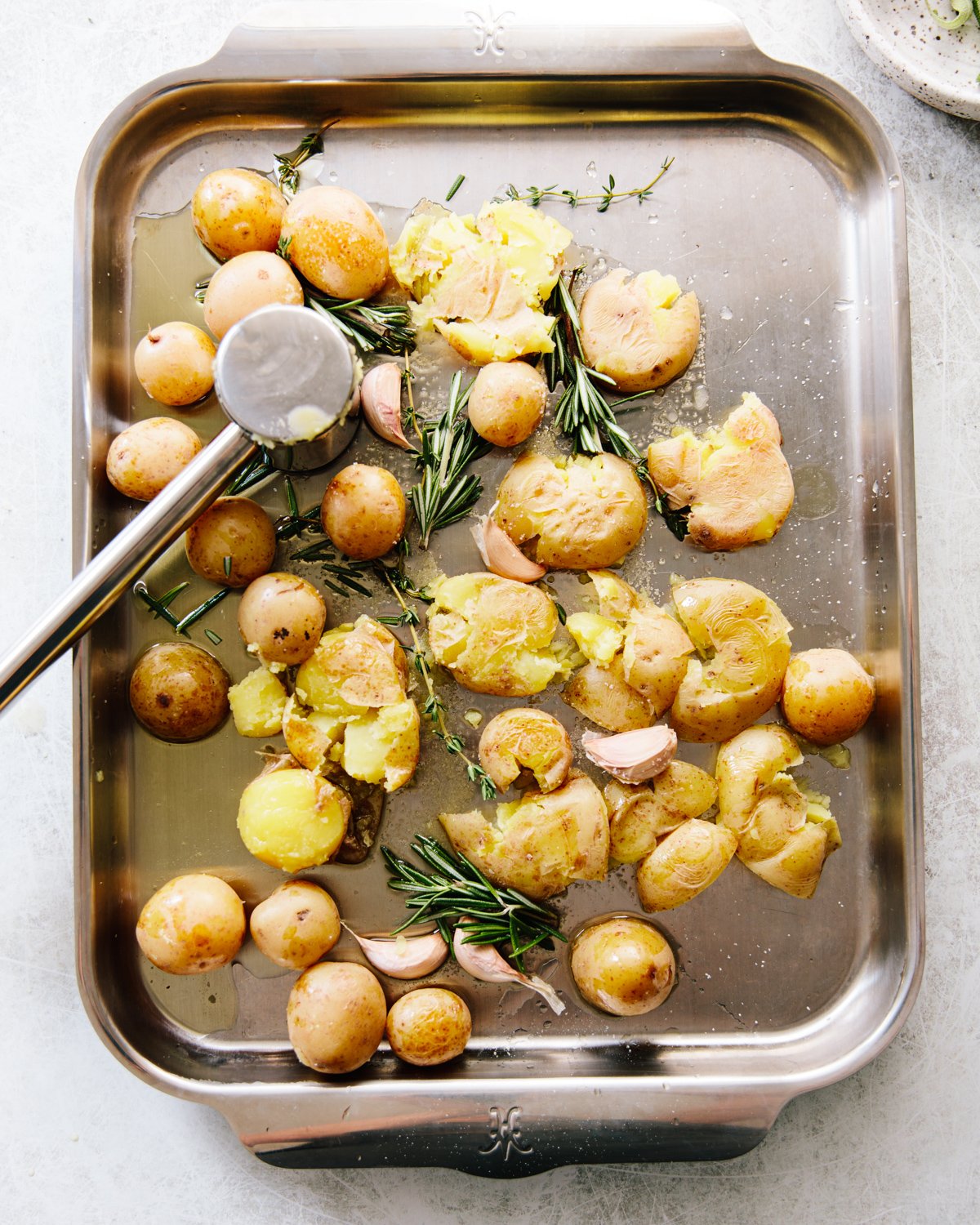 A tray of baby potatoes with sprigs of rosemary and garlic cloves, partially flattened with a metal masher on a stainless steel baking sheet.