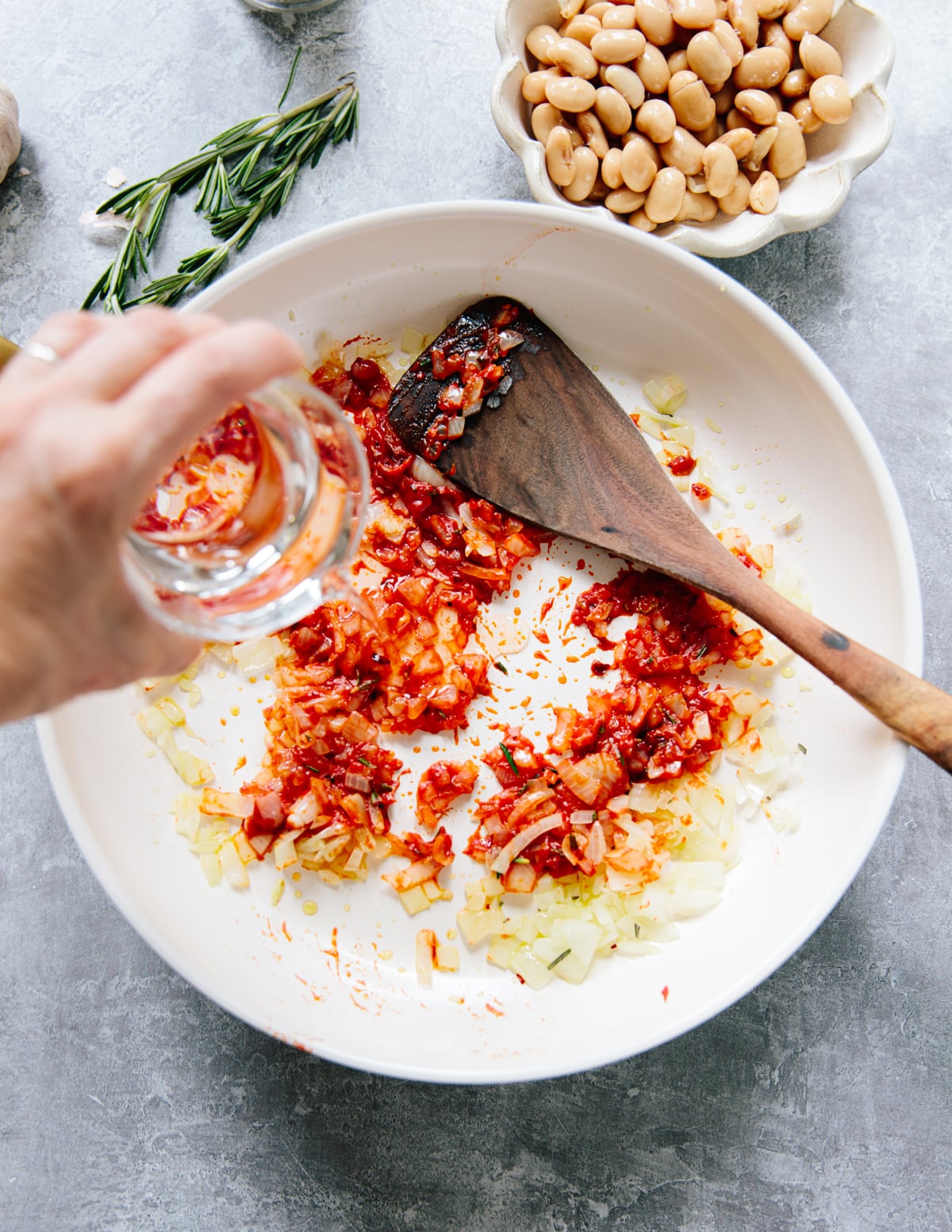 A hand pours clear vodka into a pan with sautéed onions and tomato paste, stirring with a wooden spatula. Nearby, a bowl of white beans and a sprig of rosemary are visible on the countertop.