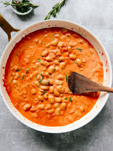 A white pan filled with creamy white beans with vodka sauce, garnished with fresh rosemary, sits on a gray surface. A wooden spoon rests inside the pan. A small bowl of chopped herbs is in the background.