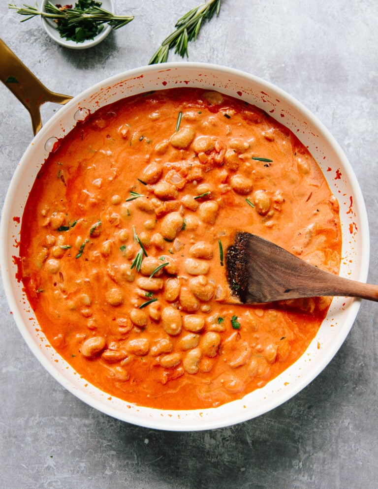 A white pan filled with creamy white beans with vodka sauce, garnished with fresh rosemary, sits on a gray surface. A wooden spoon rests inside the pan. A small bowl of chopped herbs is in the background.