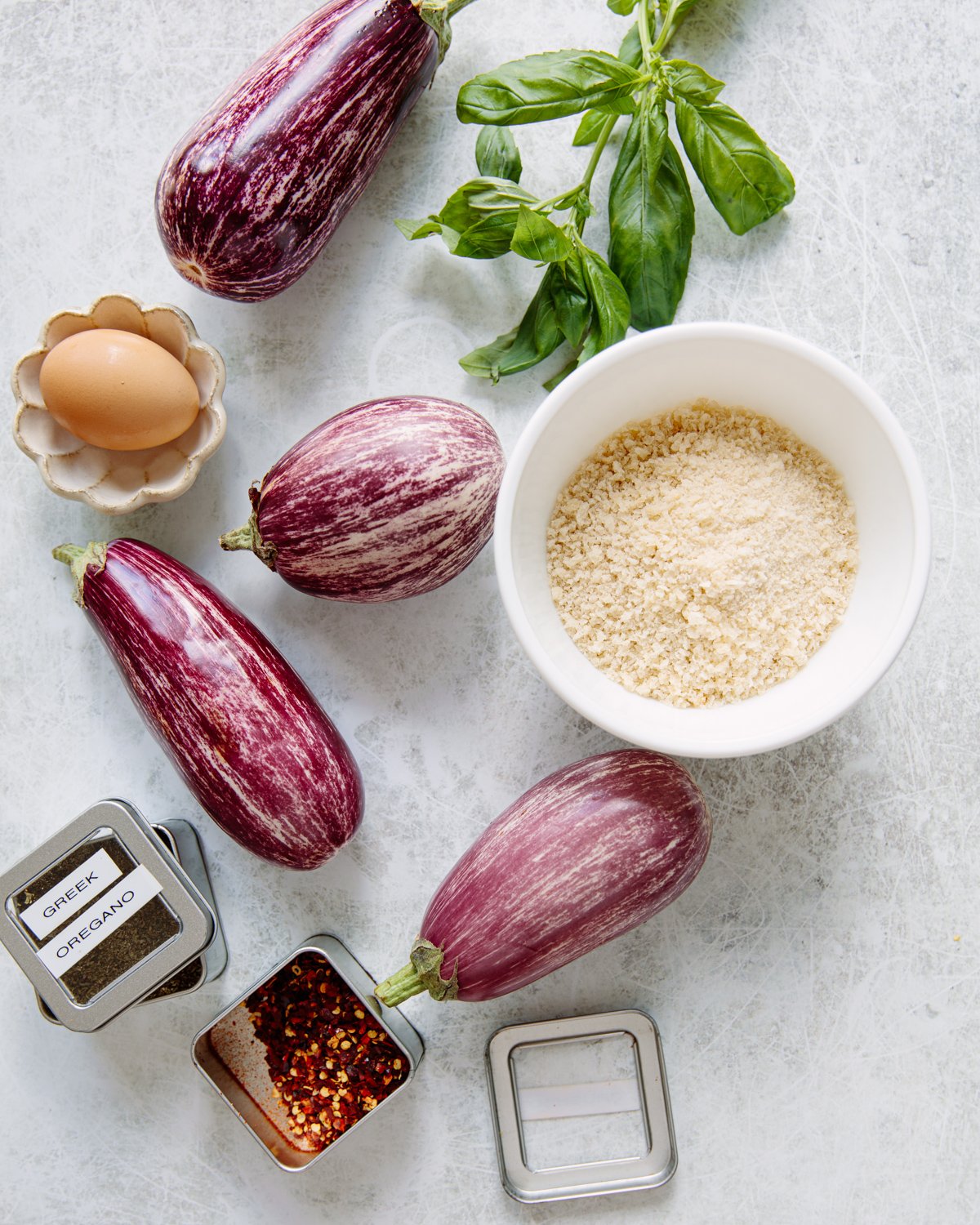 Five striped purple eggplants, a bunch of basil, a brown egg, a bowl of breadcrumbs, and small tins of oregano and crushed red pepper flakes are arranged on a light surface.