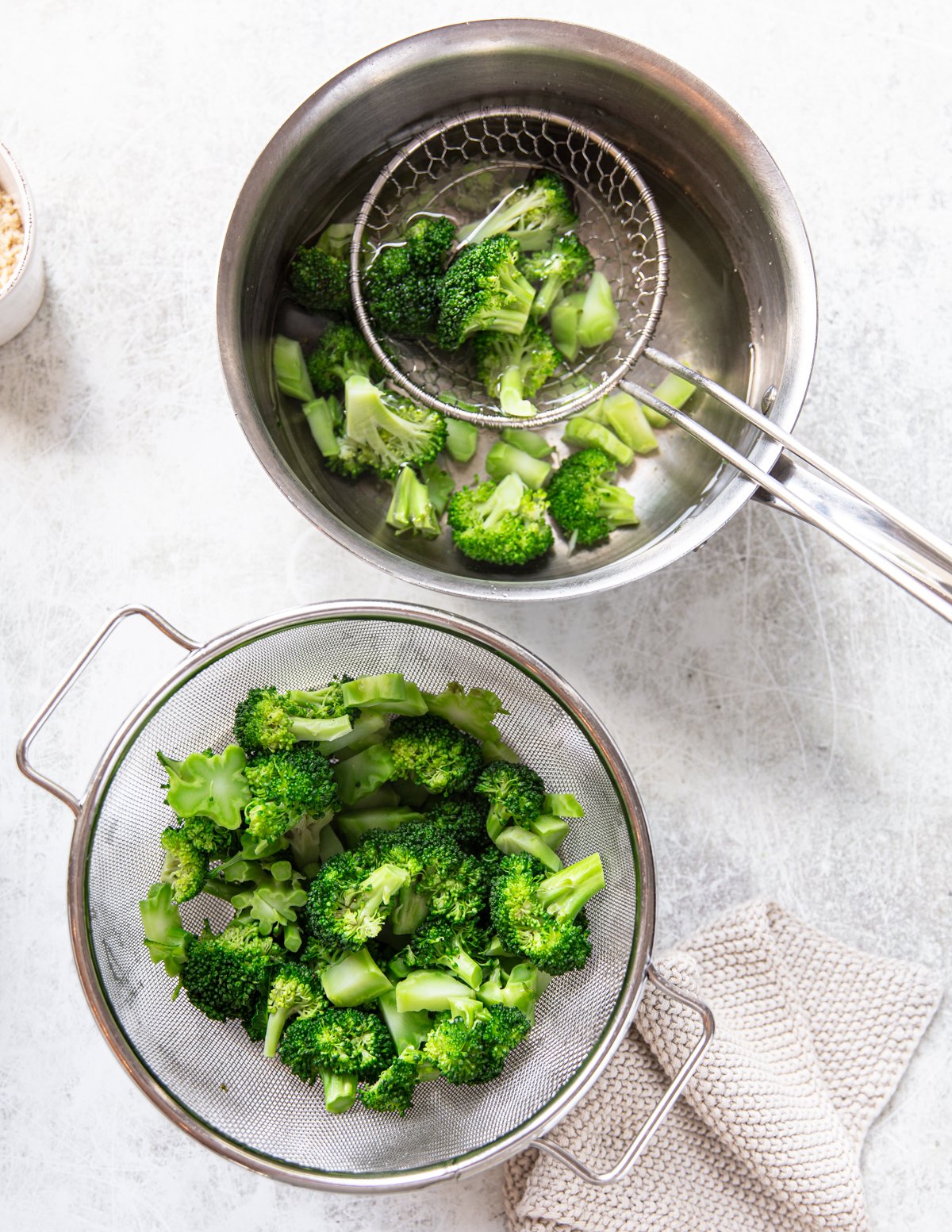 Broccoli florets are being blanched in a pot of hot water and then transferred with a slotted spoon to a metal strainer, with a knitted towel and bowl nearby on a light countertop.