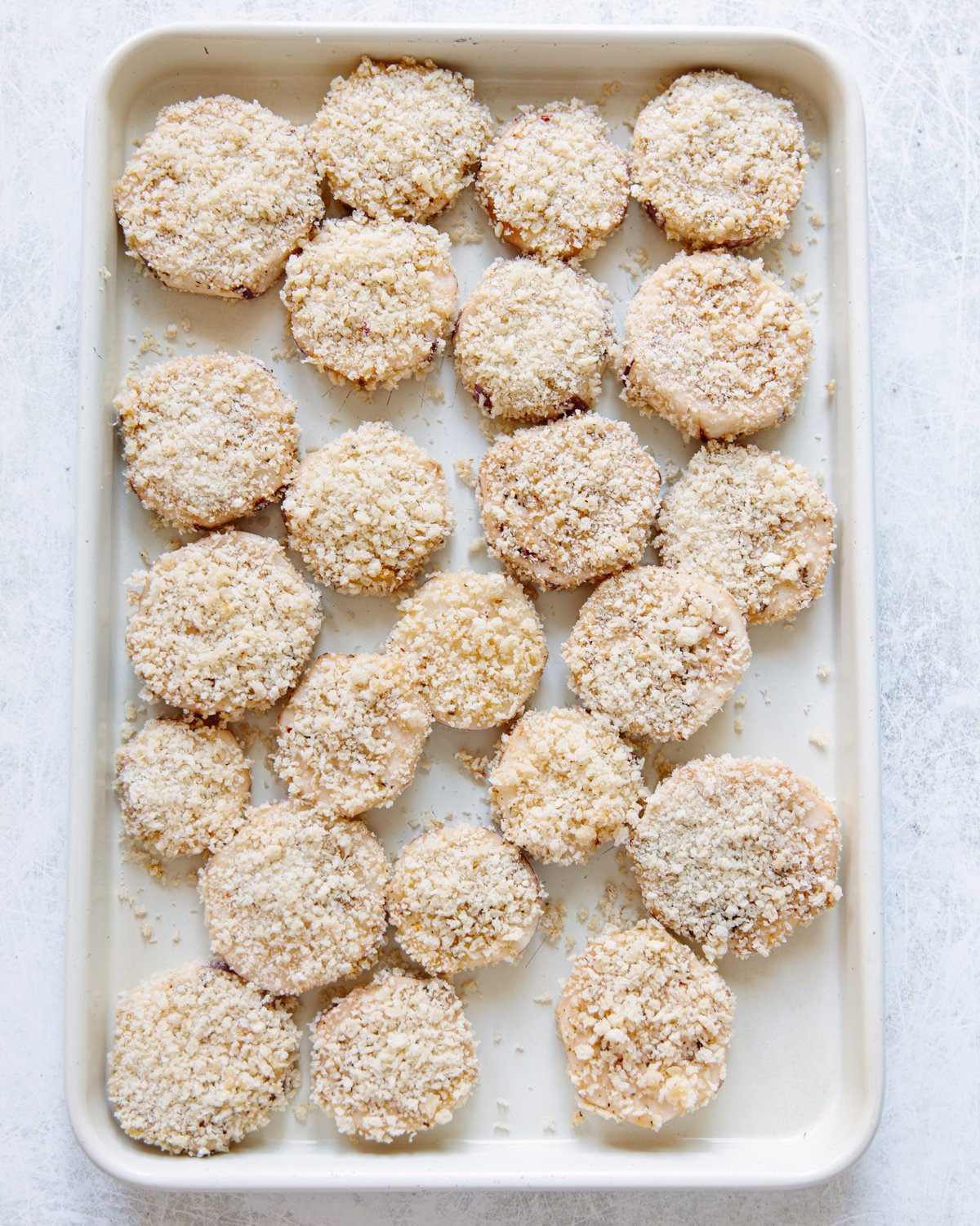 A baking sheet filled with evenly spaced, round, breaded pieces of eggplant slices, ready to be baked. The pieces are coated in a light, crumbly topping, set on a white surface.