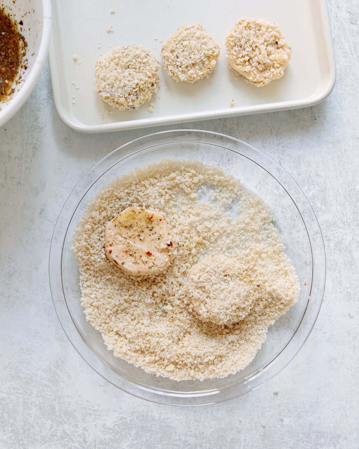 Round slices of eggplant are being coated in panko crumbs in a glass dish, with more finished pieces on a tray above on a white surface.