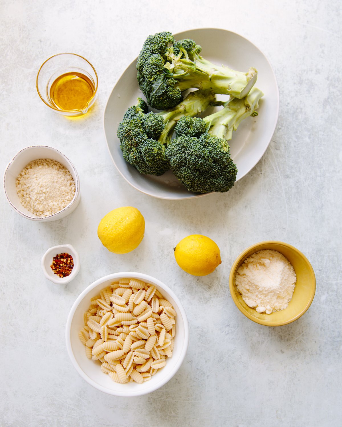 A white surface with ingredients arranged for a recipe: Fresh broccoli on a plate, two lemons, dried cavatelli pasta in a bowl, grated cheese in a yellow bowl, olive oil in a glass cup, red pepper flakes, and breadcrumbs.