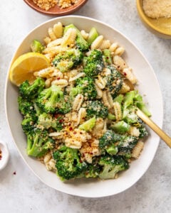 A bowl of cavatelli with broccoli florets, topped with grated cheese, breadcrumbs, and red pepper flakes. A lemon wedge sits on the side, a gold fork rests in the bowl, and small bowls with cheese and seasonings surround the dish.