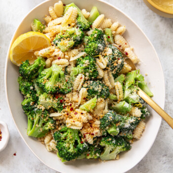 A bowl of cavatelli with broccoli florets, topped with grated cheese, breadcrumbs, and red pepper flakes. A lemon wedge sits on the side, a gold fork rests in the bowl, and small bowls with cheese and seasonings surround the dish.