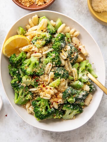 A bowl of cavatelli with broccoli florets, topped with grated cheese, breadcrumbs, and red pepper flakes. A lemon wedge sits on the side, a gold fork rests in the bowl, and small bowls with cheese and seasonings surround the dish.