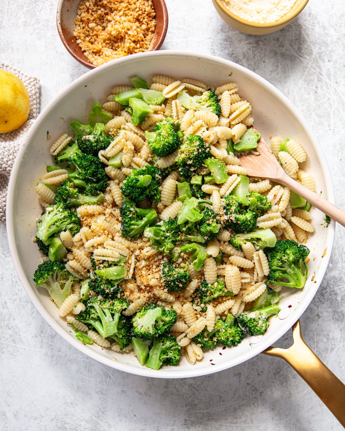 A skillet filled with cavatelli pasta and broccoli, topped with breadcrumbs. A wooden spoon rests inside, and small bowls with grated cheese and breadcrumbs are nearby on a light-colored surface.