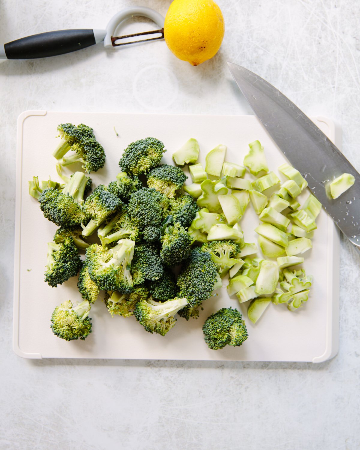 A cutting board with broccoli florets and chopped broccoli stems, a knife with broccoli bits on the blade, a vegetable peeler, and a lemon on a light-colored surface.