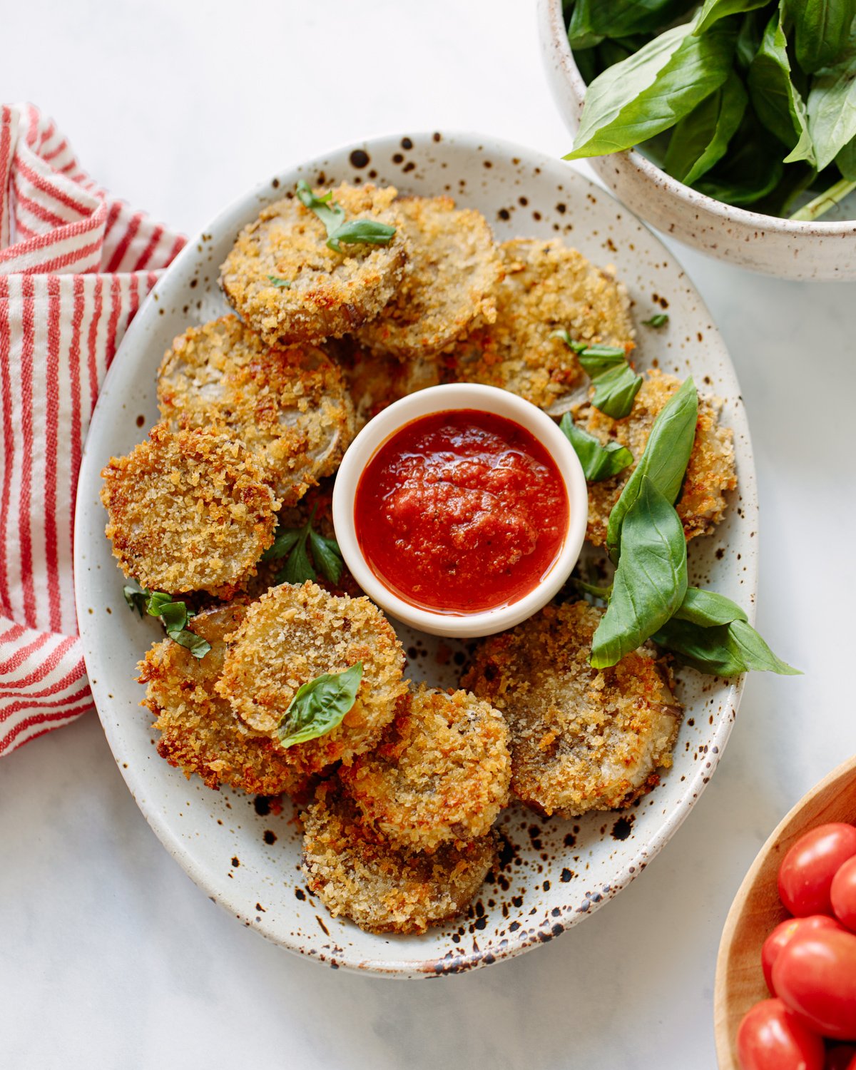 A plate of crispy breaded fried eggplant rounds garnished with fresh basil leaves, served with a small bowl of marinara sauce. A striped red towel and a bowl of basil leaves are nearby.