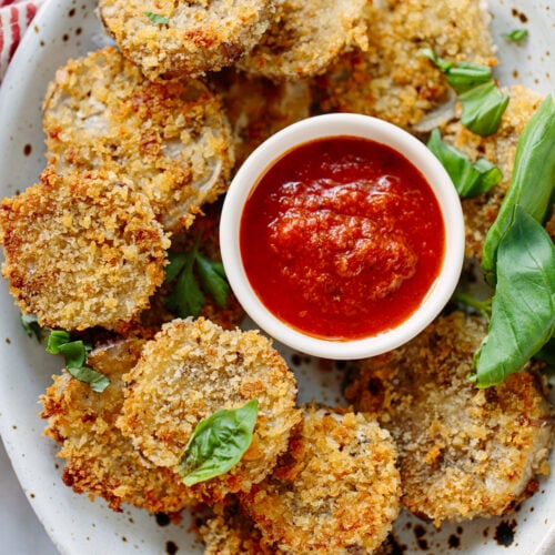 A plate of crispy breaded fried eggplant rounds garnished with fresh basil leaves, served with a small bowl of red marinara sauce for dipping.