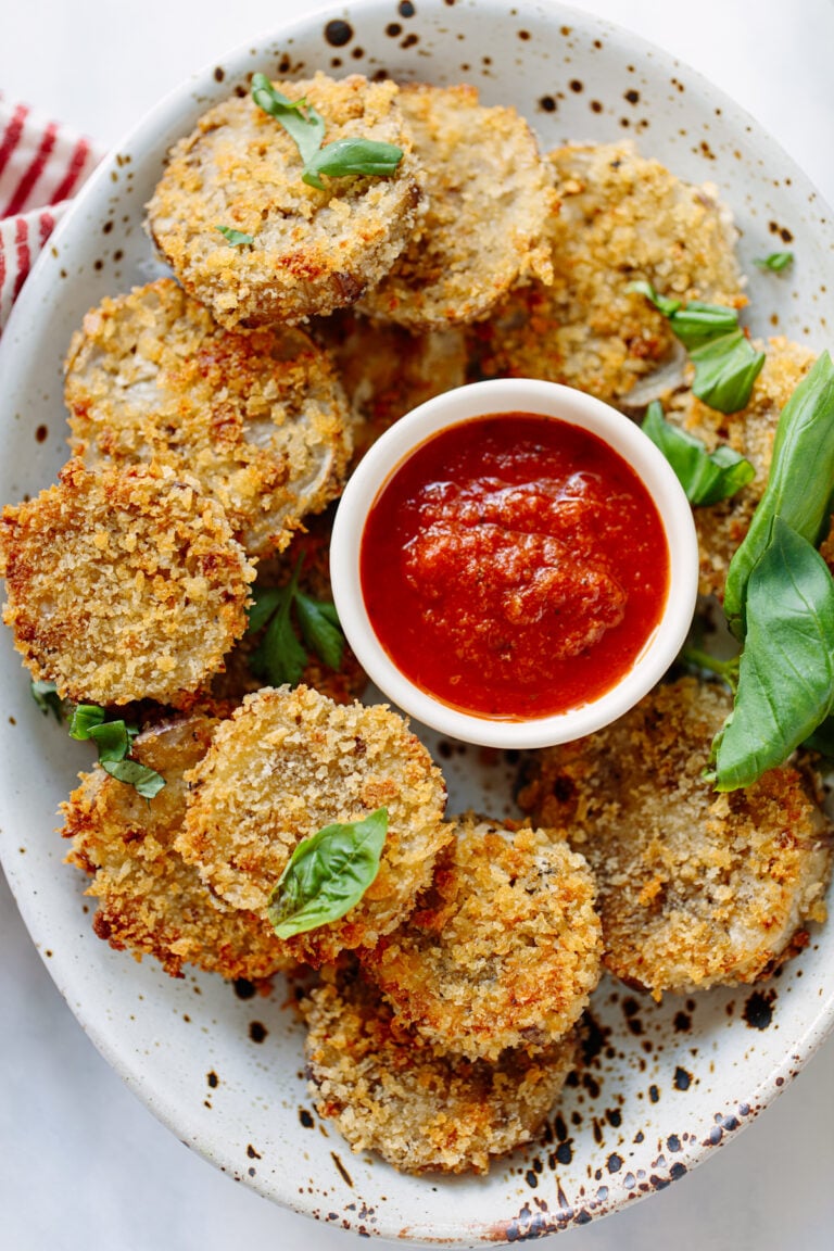 A plate of crispy breaded fried eggplant rounds garnished with fresh basil leaves, served with a small bowl of red marinara sauce for dipping.