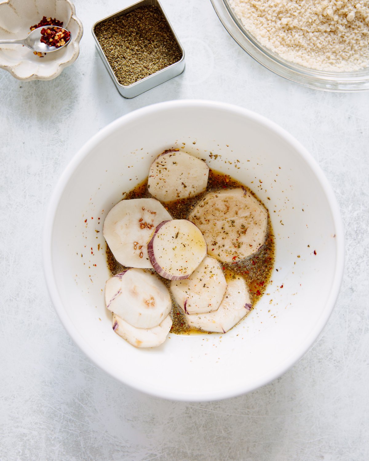 A white bowl contains sliced eggplant marinating in a seasoned egg wash, surrounded by a dish of red pepper flakes, a square container of dried herbs, and a bowl of breadcrumbs on a light surface.