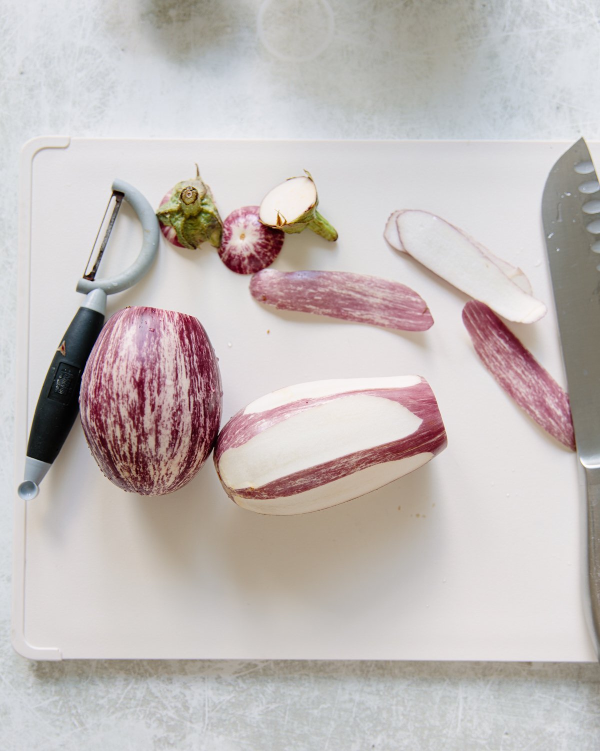 A white cutting board with a striped eggplant being peeled, a vegetable peeler, a knife, sliced eggplant pieces, and eggplant tops.