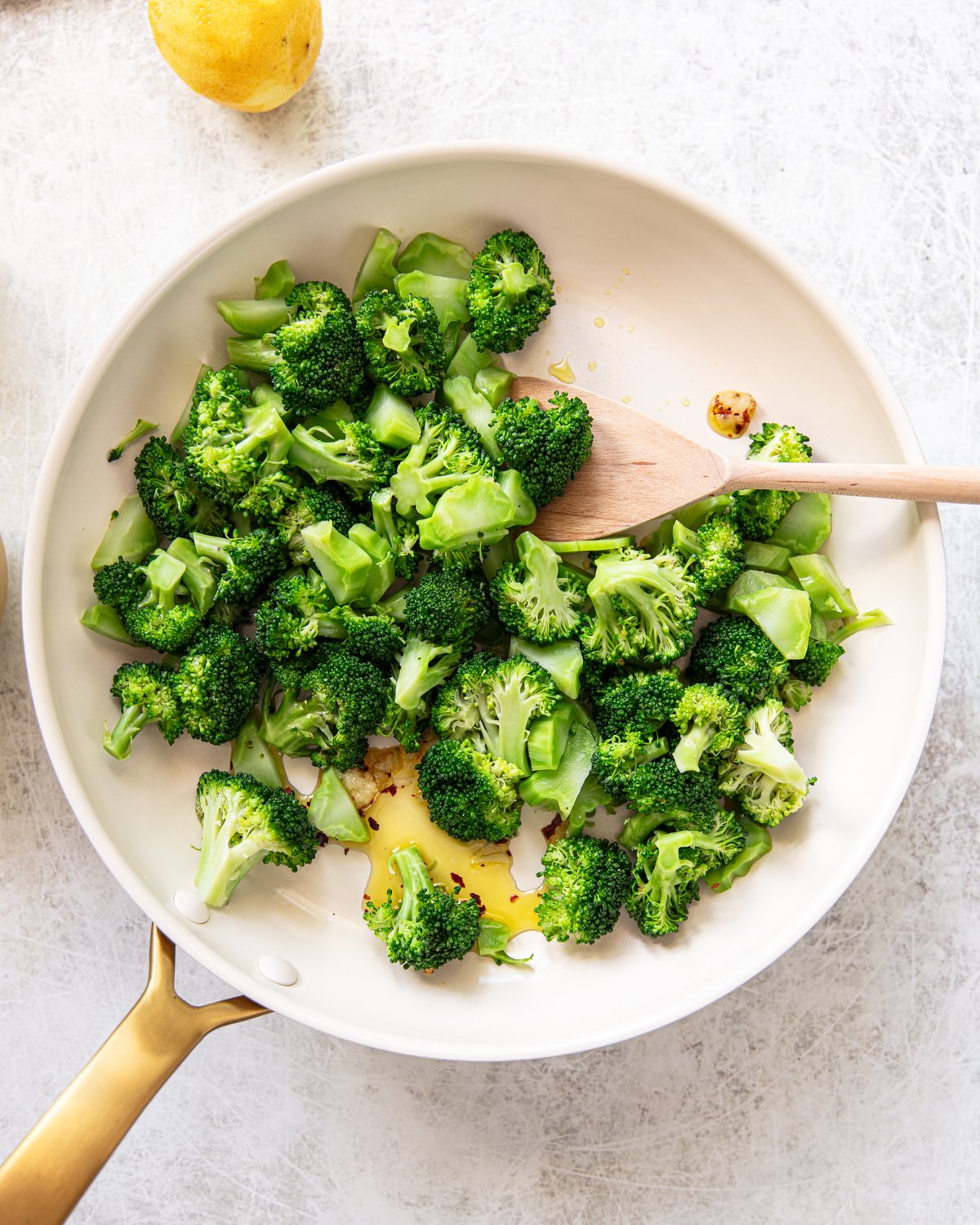 A white skillet with sautéed broccoli florets and a wooden spatula on a light surface. A lemon is partially visible in the top left corner.