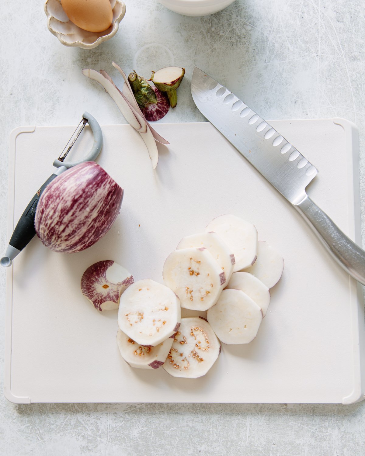 A cutting board with a partially peeled eggplant, round slices of eggplant, a chefs knife, a vegetable peeler, and an egg in a small bowl on a light countertop.