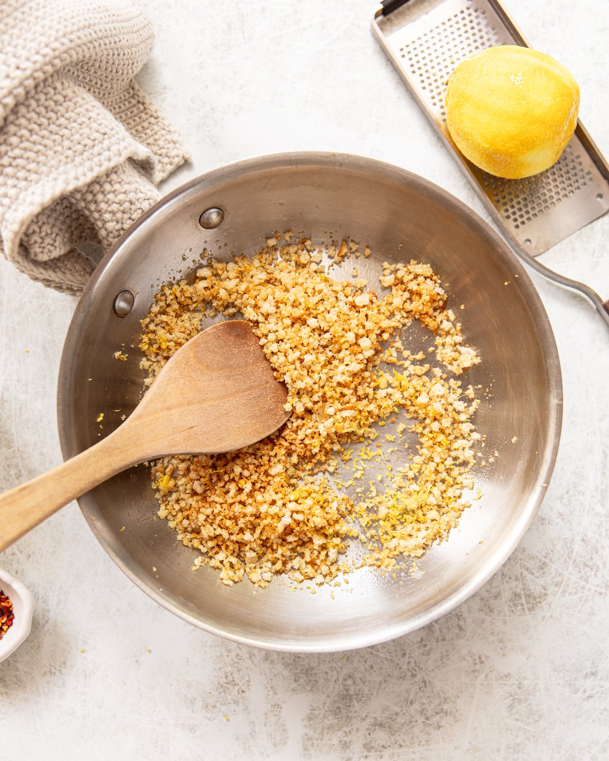 A stainless steel pan with toasted panko breadcrumbs and lemon zest being stirred with a wooden spatula. Nearby are a whole lemon, a metal grater, a beige towel, and a small bowl of red pepper flakes.